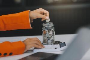 mooi Aziatisch zakenvrouw insert munten in een pin varkentje bank Aan de tafel. besparing geld, investering, valuta concept foto