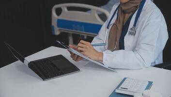 een professioneel en gefocust Aziatisch vrouw dokter in scrubs is werken en lezing medisch Onderzoek Aan haar laptop in haar kantoor Bij een ziekenhuis. foto