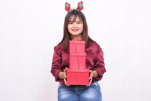 mooi jong zuidoosten Aziatisch vrouw is vrolijk terwijl draag- 3 dozen van cadeaus Bij Kerstmis vervelend een de kerstman claus hoofdband en een modern rood overhemd Aan een wit achtergrond voor Promotie en reclame foto