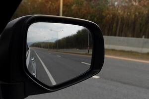 visie van spiegels vleugel. achterzijde visie van een grijs auto met asfalt weg en groen bomen en helder zonlicht Bij dag. foto