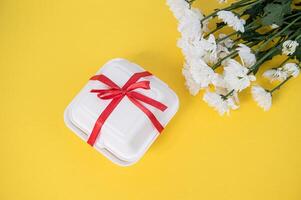wit geschenk doos met rood lint en wit chrysant bloemen Aan een geel tafel foto