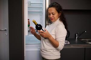gelukkig Latijns Amerikaans jong vrouw kiezen wijn, staand Bij Open koelkast in de huis keuken. perfect avondeten met perfect wijn. mensen. levensstijl. huiselijk leven. foto
