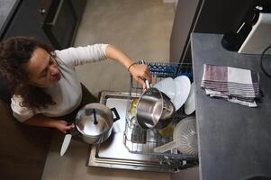 visie van bovenstaand van een jong mooi vrouw, huisvrouw aan het doen huishouden werk in de huis keuken, bezig met laden vuil borden, leeg uit of lossen vaatwasser met gebruiksvoorwerpen. keuken huishoudelijke apparaten, levensstijl visie foto