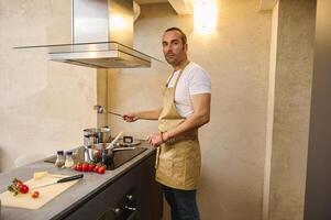 Kaukasisch knap jong Mens chef jaren 40, gekleed in beige schort, staand Bij keuken balie, Koken heerlijk avondeten voor zijn familie, glimlachen en vol vertrouwen op zoek Bij camera. ingrediënten Aan de tafel foto