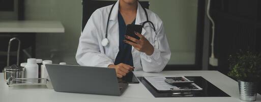 echt vrouw dokter gebruik makend van laptop en schrijven aantekeningen in medisch logboek zittend Bij bureau. jong vrouw professioneel medisch arts vervelend wit jas en stethoscoop werken Aan computer Bij werkplek. foto