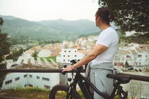 achterzijde visie van een Europese mannetje fietser ontspannende Aan top van berg, houden handen Aan stuur van zijn elektrisch trapondersteuning fiets, genieten van verbazingwekkend stedelijk landschap onderstaand. reizen en actief levensstijl foto