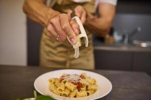 focus Aan handen van een mannetje chef in een beige schort, rooster Parmezaanse kaas kaas in een schotel van spaghetti met tomaat saus, Koken avondeten volgens naar traditioneel Italiaans recept in de huis keuken. foto