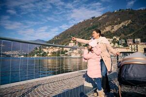 jong vrouw, moeder van twee schattig kinderen wandelen met haar dochter en pasgeboren baby slapen in de baby kinderwagen, wandelen langs de meer van komen, bewonderend de schoonheid van natuur. Italië, como, Milaan, Lombardije foto