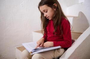 Kaukasisch schattig kind, elementair leeftijd slim weinig kind meisje bladeren door de Pagina's van een hardcover boek, lezing een boek, zittend Aan de stappen Bij huis, over- wit muur achtergrond van een landelijk huis. foto