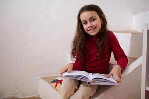 authentiek portret van een aanbiddelijk kind meisje Holding boek, glimlachen op zoek Bij camera, zittend Aan stappen Bij huis foto