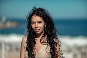 zee vrouw rust uit. portret van een vrouw met lang gekruld zwart haar- in een beige jurk staat Aan een balkon tegen de backdrop van de zee. toerist reis naar de zee. foto