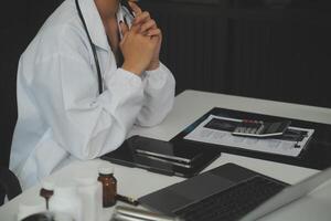 echt vrouw dokter gebruik makend van laptop en schrijven aantekeningen in medisch logboek zittend Bij bureau. jong vrouw professioneel medisch arts vervelend wit jas en stethoscoop werken Aan computer Bij werkplek. foto