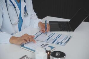 een professioneel en gefocust Aziatisch vrouw dokter in scrubs is werken en lezing medisch Onderzoek Aan haar laptop in haar kantoor Bij een ziekenhuis. foto