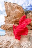 een vrouw in een rood zijde jurk staat door de oceaan, met bergen in de achtergrond, net zo haar jurk zwaait in de wind. foto