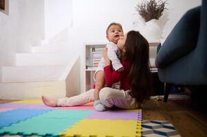 aanbiddelijk weinig kind meisje zittend Aan kleurrijk puzzel tapijt, Holding en zoenen haar weinig broer, spelen met een schattig baby jongen in licht interieur van knus landelijk huis. familie relaties concept foto