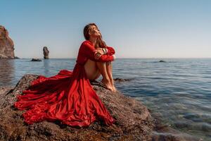 mooi sensueel vrouw in een vliegend rood jurk en lang haar, zittend Aan een rots bovenstaand de mooi zee in een groot baai. foto