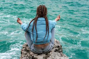 een vrouw in een blauw jasje zit Aan een rots bovenstaand een klif bovenstaand de zee, op zoek Bij de stormachtig oceaan. meisje reiziger rust, denkt na, dromen, geniet natuur. vrede en kalmte landschap, winderig het weer. foto