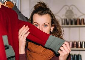 een vrolijk vrouw met gekruld haar- speels verbergt achter een rood kledingstuk, tonen haar ogen in een winkel instelling met een knus backdrop foto