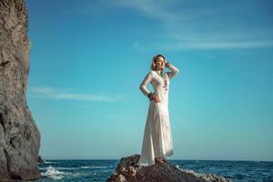 vrouw wit jurk zee stenen rotsen.van middelbare leeftijd vrouw looks mooi zo met blond haar, boho stijl in een wit lang jurk Aan strand sieraden in de omgeving van haar nek en armen. foto