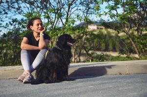 multi etnisch mooi nadenkend vrouw zittend De volgende naar haar hond Aan de asfalt, een mooi zwart cocker spaniel en op zoek dromerig in de afstand. huisdieren en mensen. vrouw wandelen haar hond Aan riem foto