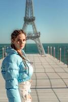 groot model- van de eiffel toren Aan de strand. een vrouw wandelingen langs de pier naar de toren, vervelend een blauw jasje en wit jeans. foto