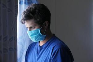 een ziek Mens met een masker Aan zijn gezicht op zoek door venster foto
