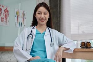 portret van mooie vrouwelijke arts van Aziatische etniciteit in uniform met stethoscoop, glimlachend en kijkend naar de camera in de kliniek van het ziekenhuis. één persoon met expertise in professionele behandeling. foto