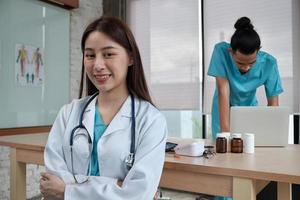 portret van mooie vrouwelijke arts van Aziatische etniciteit in uniform met stethoscoop. glimlach en kijk naar de camera in een ziekenhuiskliniek, mannelijke partner die achter haar werkt, twee professionele personen.