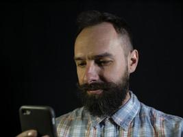 stijlvolle man met een baard en snor kijkt zorgvuldig naar de telefoon op een zwarte achtergrond foto