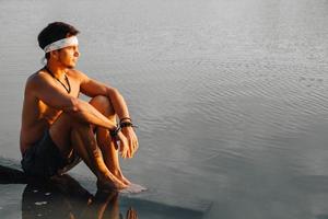 man met perfect gespierde zittend op houten brug en kijkt naar de waterreflex bij zonsondergang foto