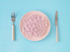 creatief lay-out gemaakt met roze bord, ijs kubussen, mes en vork Aan wit en blauw achtergrond. minimaal nul calorie voedsel concept. modieus gezond voedsel met laag calorieën grappig idee. vlak leggen. foto