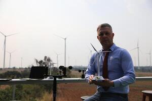 een senior mannetje energie specialist is inspecteren wind turbines Aan een ecologisch bouw project gebruik makend van een wind snelheid meten instrument Bij een windmolen. foto