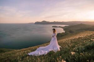 een vrouw in een wit jurk staat Aan een heuvel met uitzicht de oceaan foto