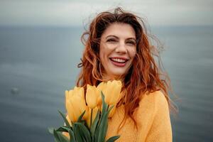 portret van een gelukkig vrouw met haar- vliegend in de wind tegen de backdrop van bergen en zee. Holding een boeket van geel tulpen in haar handen, vervelend een geel trui foto