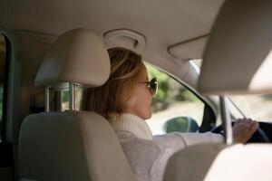 een blond vrouw in een wit trui en jeans is het rijden. gelukkig vrouw zittend in een auto met een wit interieur. foto