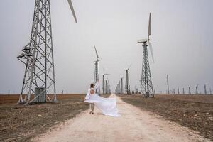 een vrouw in een wit jurk is wandelen naar beneden een aarde weg in voorkant van een rij van wind turbines. foto