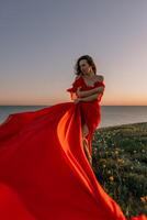 vrouw rood jurk staand met gras begroeid heuvel. de zon is instelling in de achtergrond, gieten een warm gloed over- de tafereel. de vrouw is genieten van de mooi visie en de vredig atmosfeer. foto