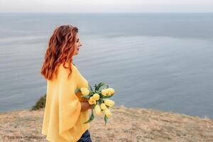 achterzijde visie van een vrouw met lang haar- tegen een achtergrond van bergen en zee. Holding een boeket van geel tulpen in haar handen, vervelend een geel trui foto