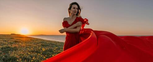 vrouw rood jurk staand met gras begroeid heuvel. de zon is instelling in de achtergrond, gieten een warm gloed over- de tafereel. de vrouw is genieten van de mooi visie en de vredig atmosfeer. foto