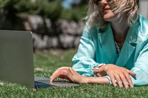 een jong mooi vrouw met blond gekruld haar- in bril en een blauw jurk zit Aan de gras in natuur en toepassingen een laptop, foto