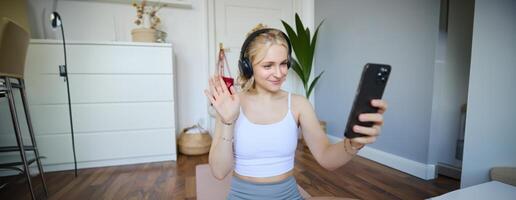 portret van jong sportief vrouw in koptelefoon, records Aan haar smartphone, leven streaming en gezegde Hallo naar volgers terwijl aan het doen training opleiding Bij huis foto