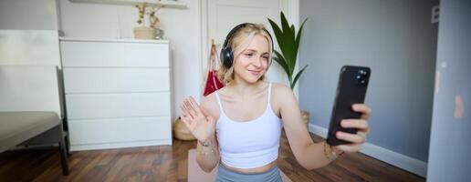 portret van jong vrouw, geschiktheid instructeur aan het doen online training sessie Aan smartphone, golvend hand- Bij camera en gezegde Hallo, zittend Aan rubber mat in een kamer foto