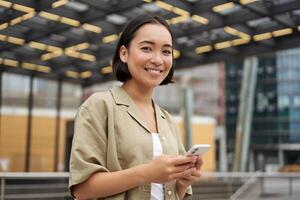 technologie mensen. jong glimlachen Aziatisch meisje Holding smartphone, gebruik makend van mobiel app terwijl staand Aan straat, navigeren in de omgeving van stad foto
