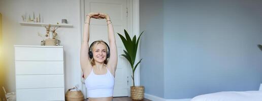 portret van vrouw aan het doen geschiktheid opdrachten Bij huis, horloges sport- Aan laptop, vervelend draadloze koptelefoon, volgt training instructies, zit Aan rubber mat in haar kamer foto