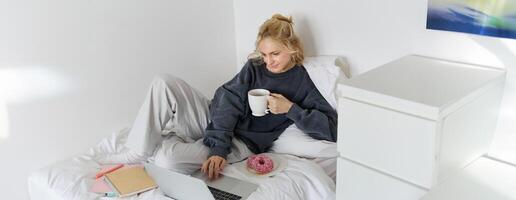 portret van jong vrouw, leerling aan het studeren in haar bed, ontspannende terwijl voorbereidingen treffen huiswerk, aan het eten donut, gebruik makend van laptop in slaapkamer en drinken thee foto