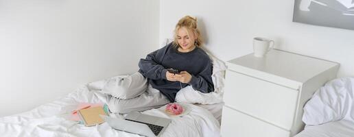 portret van glimlachen openhartig vrouw, aan het liegen in bed met donut, gebruik makend van smartphone en laptop, resting Bij huis in slaapkamer, aan het kijken TV tonen of chatten online foto