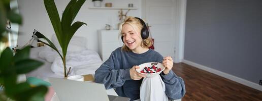 portret van glimlachen jong vrouw, aan het kijken TV tonen in koptelefoon, aan het eten ontbijt en op zoek Bij laptop scherm foto