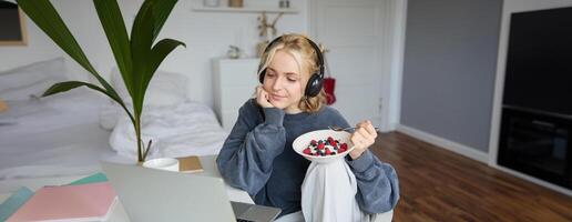 portret van gelukkig jong blond vrouw, zittend in een kamer, aan het kijken film Aan laptop en aan het eten gezond ontbijt, drinken thee, resting Aan weekend foto