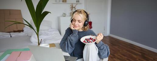 beeld van gelukkig vrouw zittend in een kamer, aan het kijken interessant TV tonen of film Aan laptop, gebruik makend van schreeuwen onderhoud, vervelend koptelefoon, aan het eten toetje en drinken thee foto