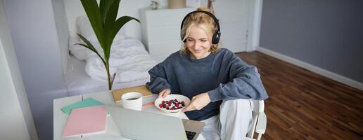 portret van glimlachen jong vrouw, aan het kijken TV tonen in koptelefoon, aan het eten ontbijt en op zoek Bij laptop scherm foto